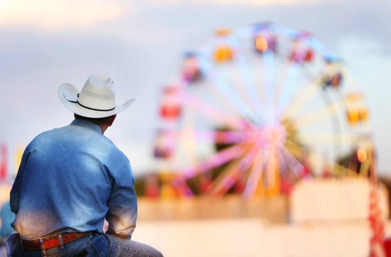 Photograph focused on a man dressed in jeans, denim shirt, cowboy hat, sitting on a bench with a blurred Ferris Wheel in the background