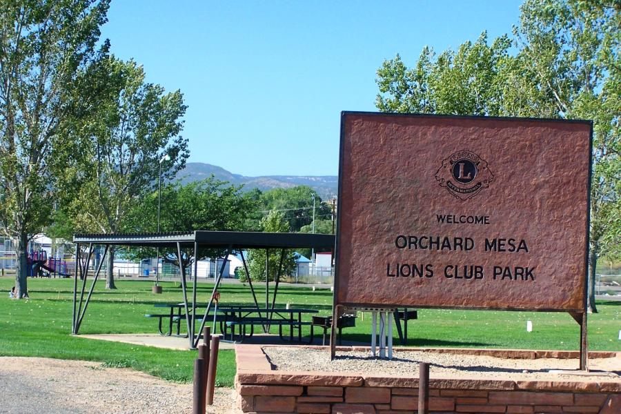 Photograph of Orchard Mesa Lions Club Park with Sign at the entrance Welcome to Orchard Mesa Lions Club Park, covered area with with picnic tables and barbeque stands, grass area, and mature trees