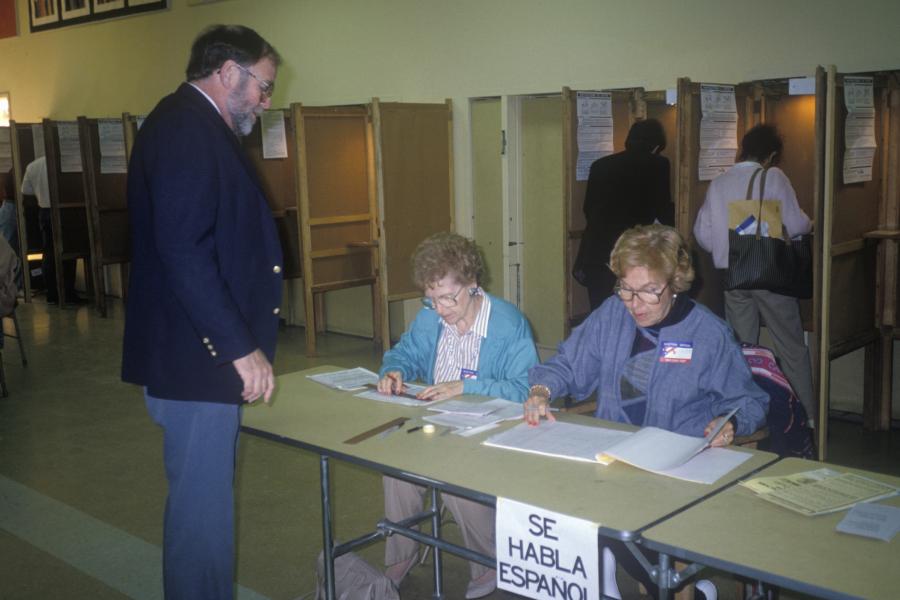 Photograph of two female volunteer working at a table assisting a man to check in at Voting Center with people in voting booths in the background