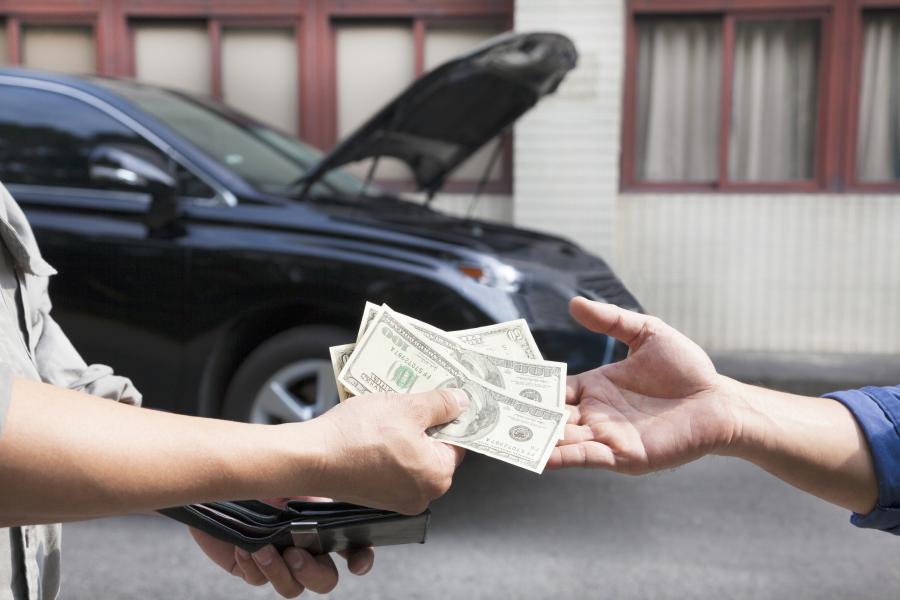 Photograph of a person selling a car, seller is handing two hundred and fifty dollars in cash to the buyer, car with hood raised in the background