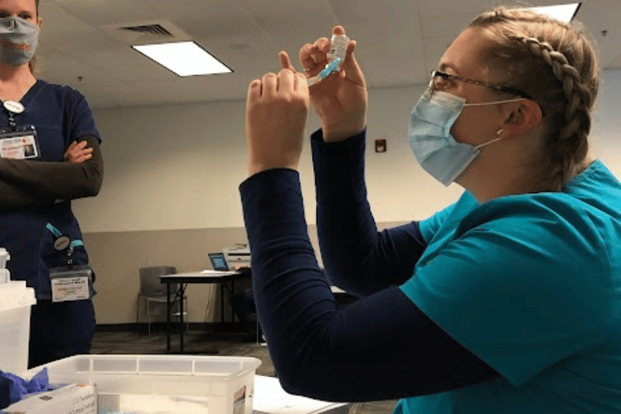 Nurse with mask on is holding a vial as she prepares a vaccine for a patient.