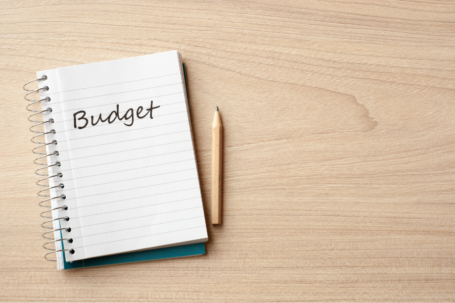 "Budget" written in notebook sitting on light tan desk next to pencil.