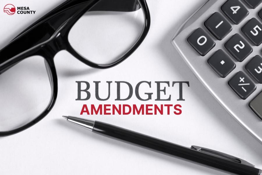 Glasses, calculator, and pen sit on white table with grey and red text reading, "Budget Amendments".