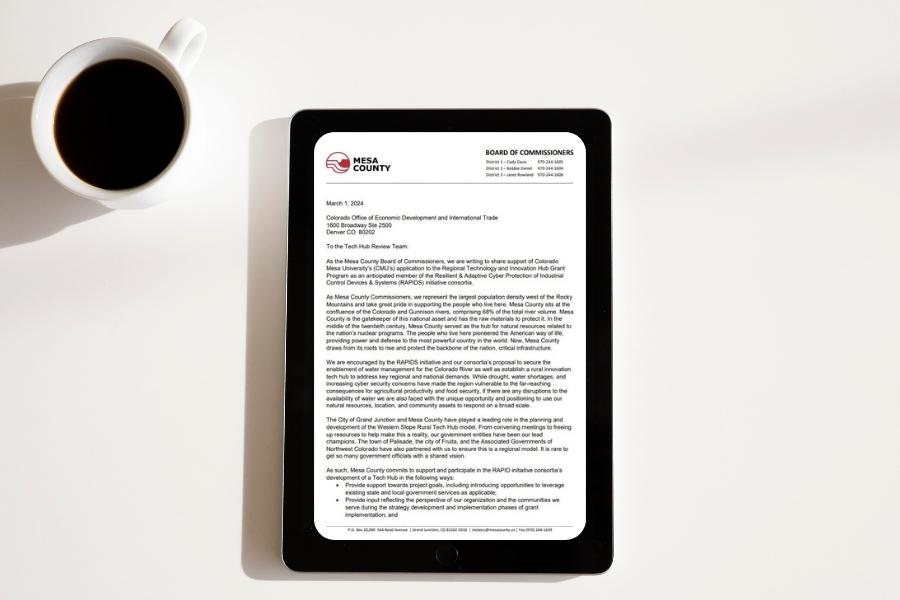 A black iPad displays a letter from Mesa County to the Office of Economic Development and International Trade next to a white mug of coffee. 