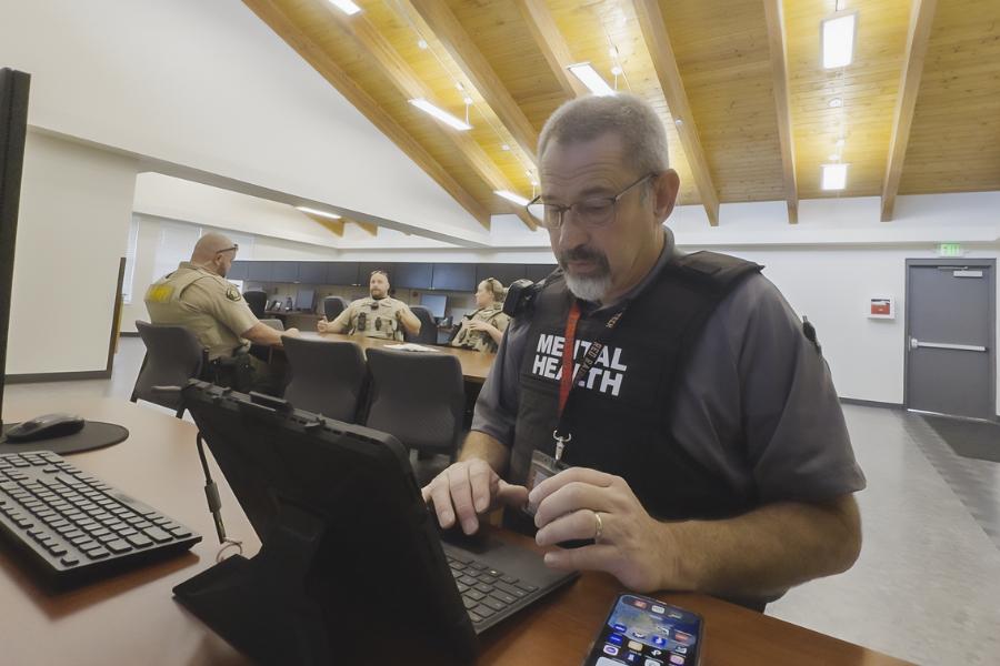 Man wearing vest that says "mental health" sits at a desk typing on a black laptop. 