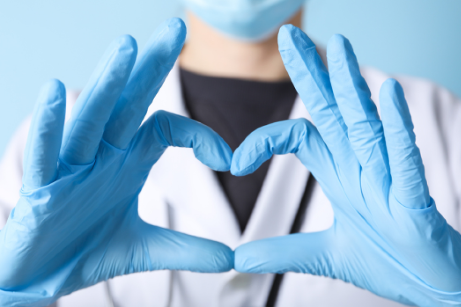Medical professional wearing a blue mask and blue, plastic gloves shaping their fingers into a heart.