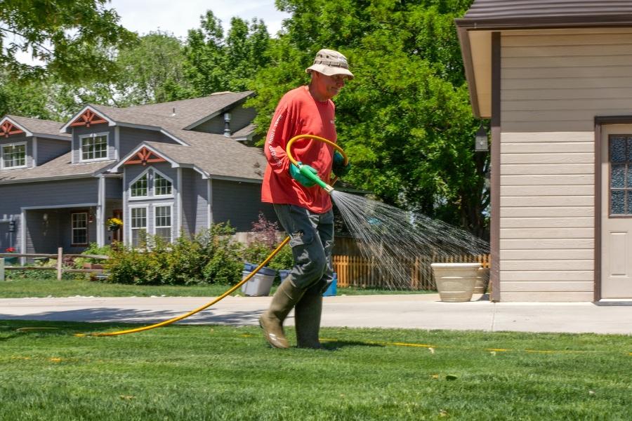 Man in red shirt and rubber boots walks along green grass spraying pesticides with a yellow hose. 