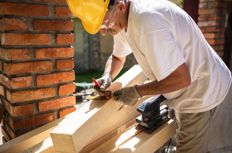 Photograph of a builder marking a 4 by 4 post for cutting with a pencil and ruler, pile of posts, and power sander