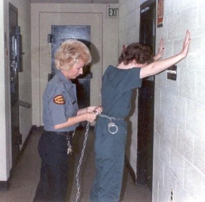 Historical photograph of a female deputy placing chain with hand cuffs on a female inmate leaning on a well with hands spread