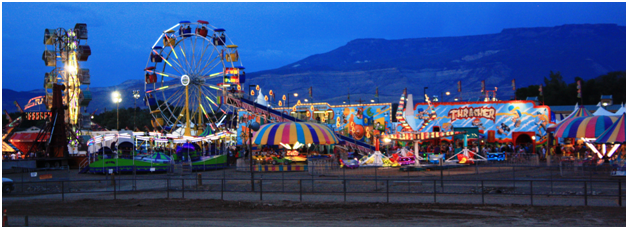 Photograph showing the Carnival Midway attractions portion of the Mesa County Fair at night.  Shows a Ferris wheel, roller coaster, merry-go-round, and other rides with a mountain range in the background