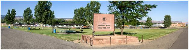 Photograph of Orchard Mesa Lions Club Park showing sign, large grass area, mature trees, covered picnic areas, road running along left and right side of park