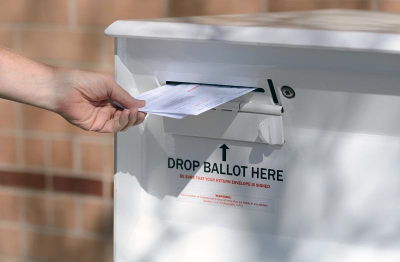 Photograph of a hand inserting two election ballots in envelops into the slot of a ballot drop box
