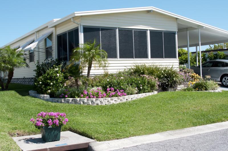 Photograph of a  Manufactured Home on lot with grass, flower bed, shrubs, palm trees, car parked under car port