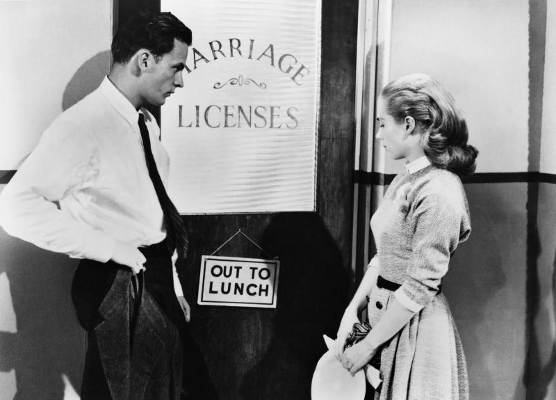 Black and White Photograph of a Couple waiting outside door with Marriage Licenses printed on the window and a "Out to Lunch" sign hanging on door knob