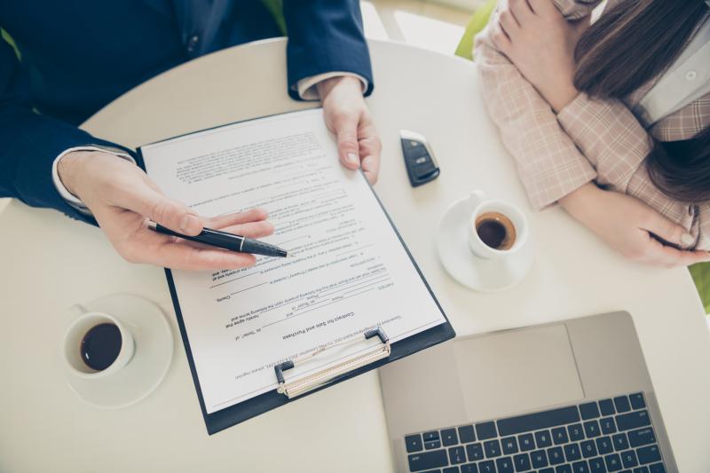 Photograph of a sales person selling a car with a contract attached to a clipboard, he is pointing with a pen to the contract, female customer sitting next to him, table has car key, laptop, and two cups of coffee