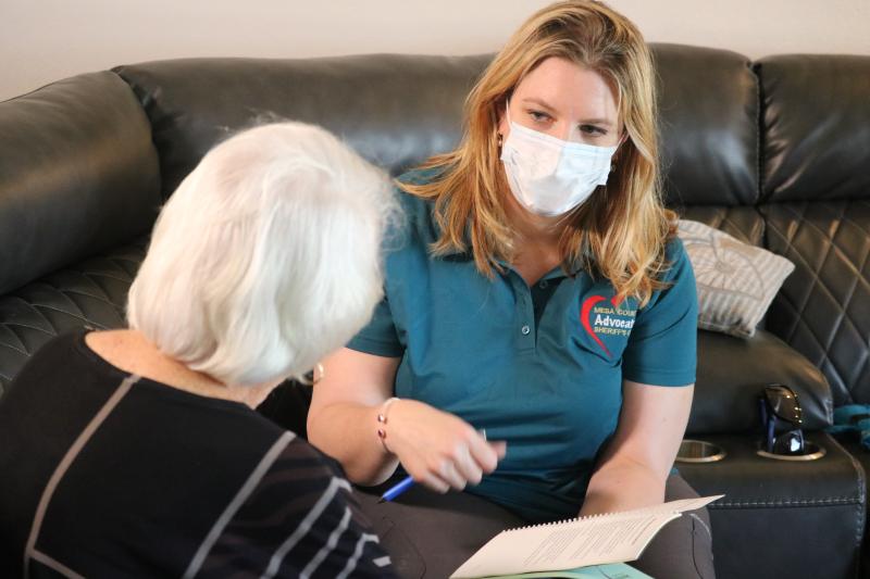 Photograph of Female Mesa County Sheriff's Office Advocate with a white mask on working with a victim complete paperwork sitting on a couch in their living room