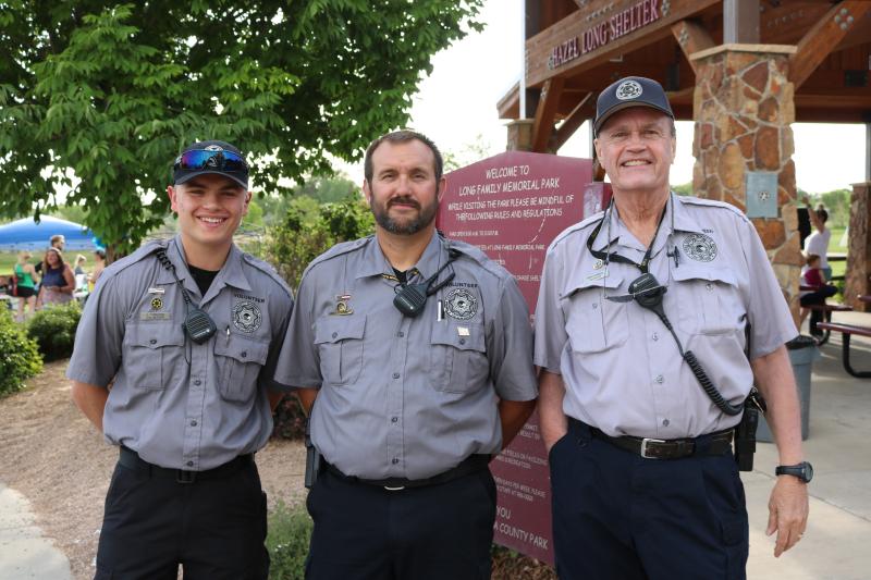 Photograph of three male Volunteer workers for the Mesa County Sheriff's Office at the Touch a truck Event held at the Long Family Park, sign and Hazel Long Shelter in the background
