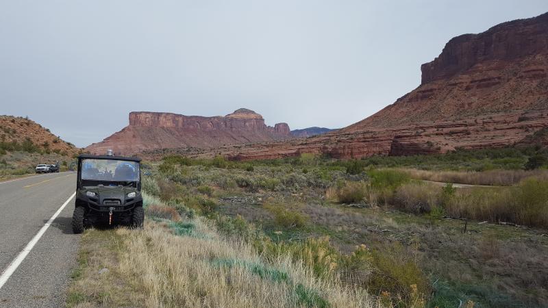 Photograph of Desert Landscape on the side of highway showing weed growth.  Jeep parked in front, mountain ranges in the background.