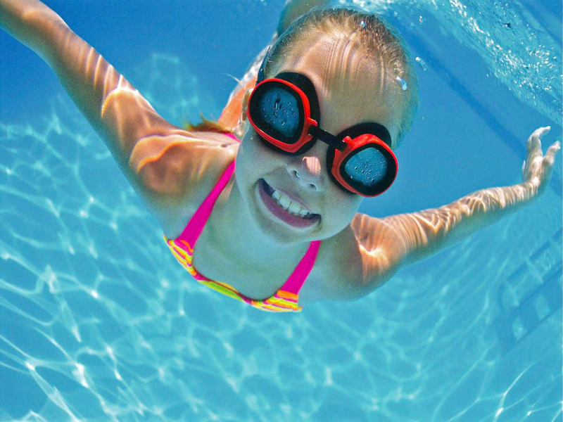 girl smiling underwater with red and black swim googles on