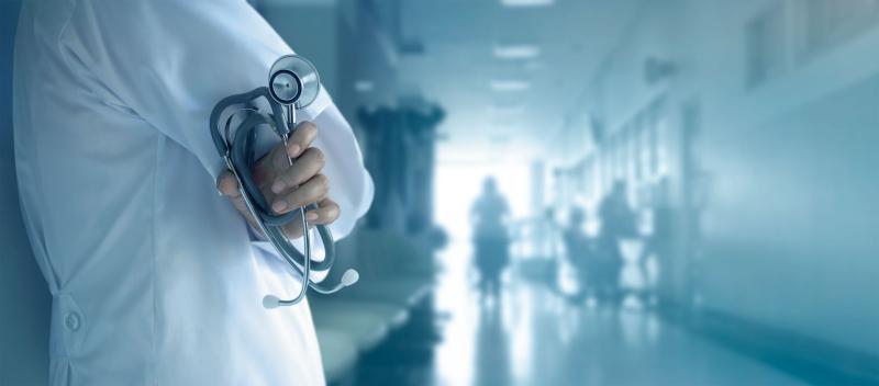 Doctor with stethoscope in front of a hospital hallway with people in the background. 
