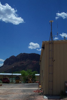 GPS survey antenna placed behind a tan metal building with dirt in the foreground, a parking lot to the side, a small rocky escarpment in the background, and a blue sky with white puffy clouds. 