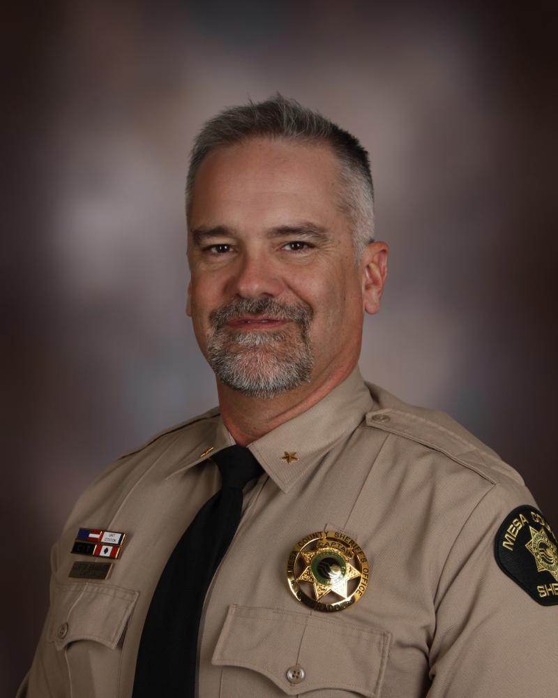 Captain Curtis Brammer Investigations and Special Teams. A white male with dark grey/brown hair sits against a grey-brown background. He has on a tan, double chest pocket shirt. He has a Mesa County Sheriff's Office badge on his left chest and his awards bars on the right. He has a slight smile with a beard and mustache.