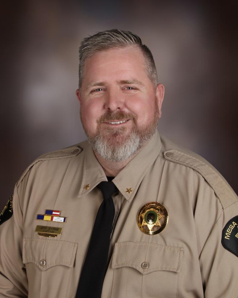Captain Jeff Byrne is a white male sitting against a grey-brown background. He has short white and grey hair with a large smile and short beard. He has on a tan shirt with chest pockets. He has a Mesa County Sheriff's Office badge on his left chest and awards bars on his right chest.