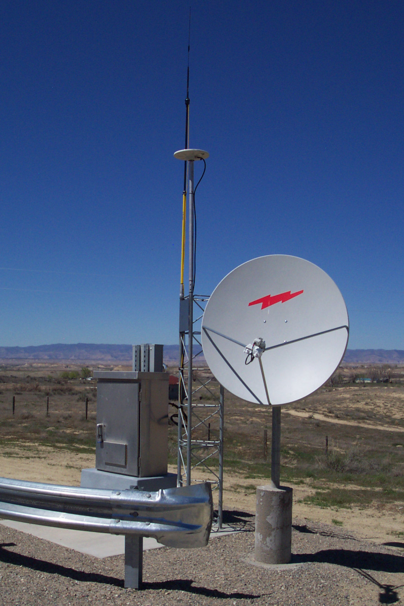 Blue sky dominating a desert landscape shows a GPS antenna set next to a small antenna dish with a car metal barrier placed in front for protection. 