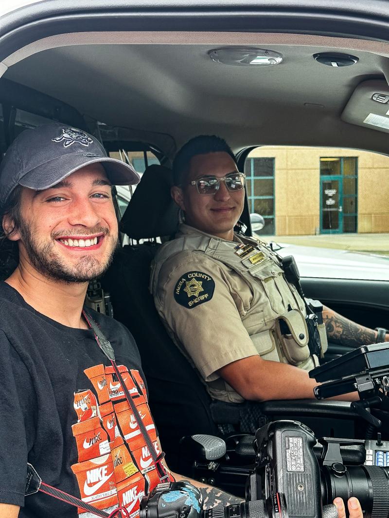 A young man and a Mesa County Sheriff's Office Deputy smile at the camera from inside of the Deputy's patrol vehicle during a ride-along.