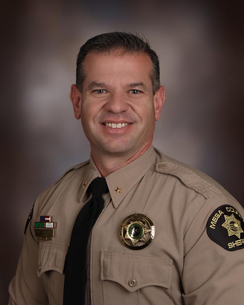 Captain Eric Sperber Patrol. A white male with dark brown/black hair wearing a tan shirt with two chest pockets sits against a brown/grey background. He has on a Mesa County Sheriff's Office badge on the left chest and his awards bards on the right chest. He has a broad smile with no facial hair.