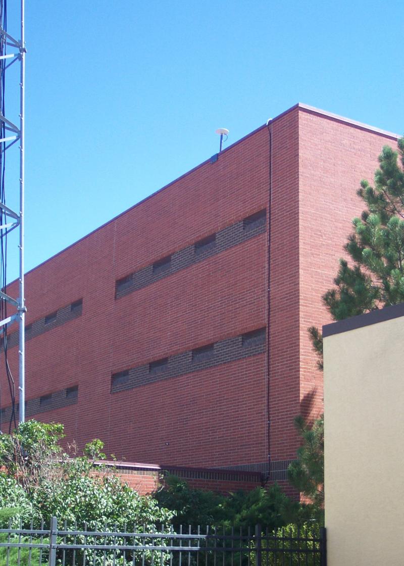 Antenna shown on top of a red brick, 3 story building, with green shrubbery and a black metal fence at the base of the building.