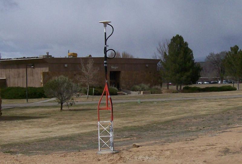 Antenna located in bare ground next to grass with a brown brick building and mature landscape in the background. 