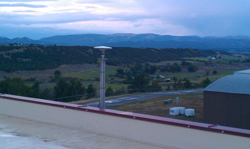 GPS antenna situated on top of a building roof with image looking out on mountains and a green valley with a blue sky. 