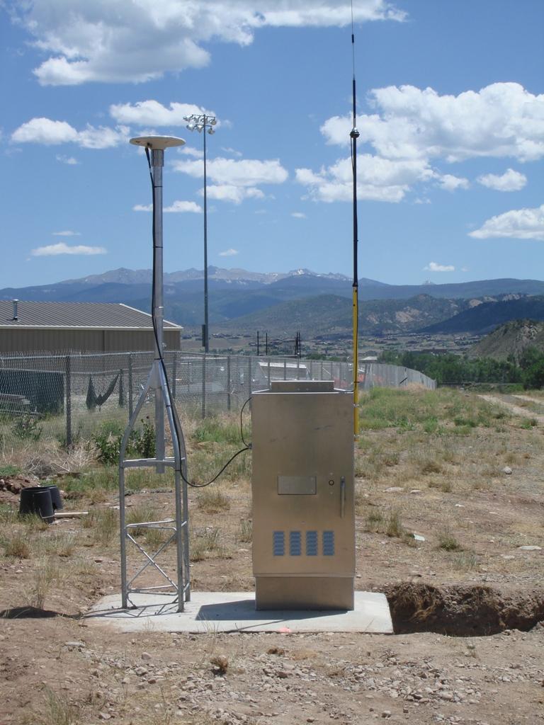 Antenna located in bare ground near a chain link fence and tan building with mountains in the background and a blue sky with fluffy white clouds. 