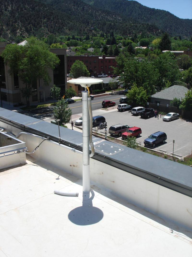 Antenna located on a white roof of multi-story building overlooking a parking lot and part of the town with green covered mountains and a blue sky in the background.