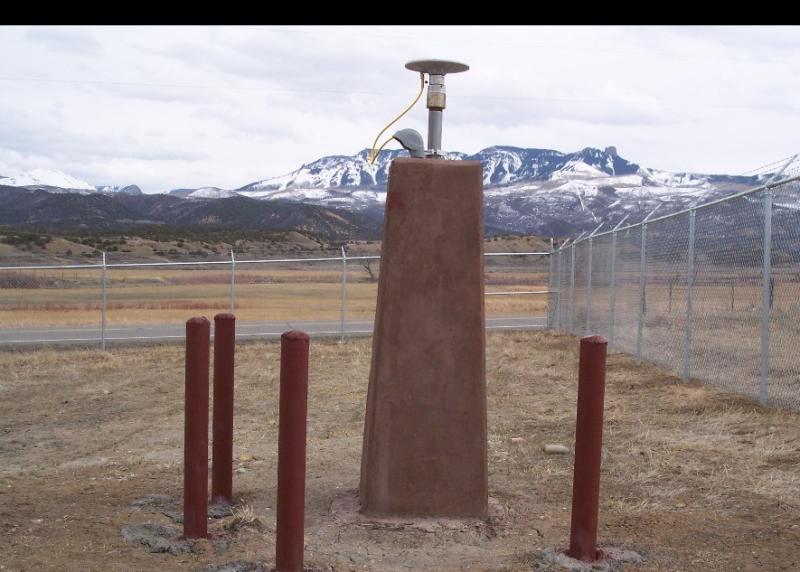Antenna located on tall, tan concrete block with 4 red-brown posts surrounding it and snow-covered mountains in the background.