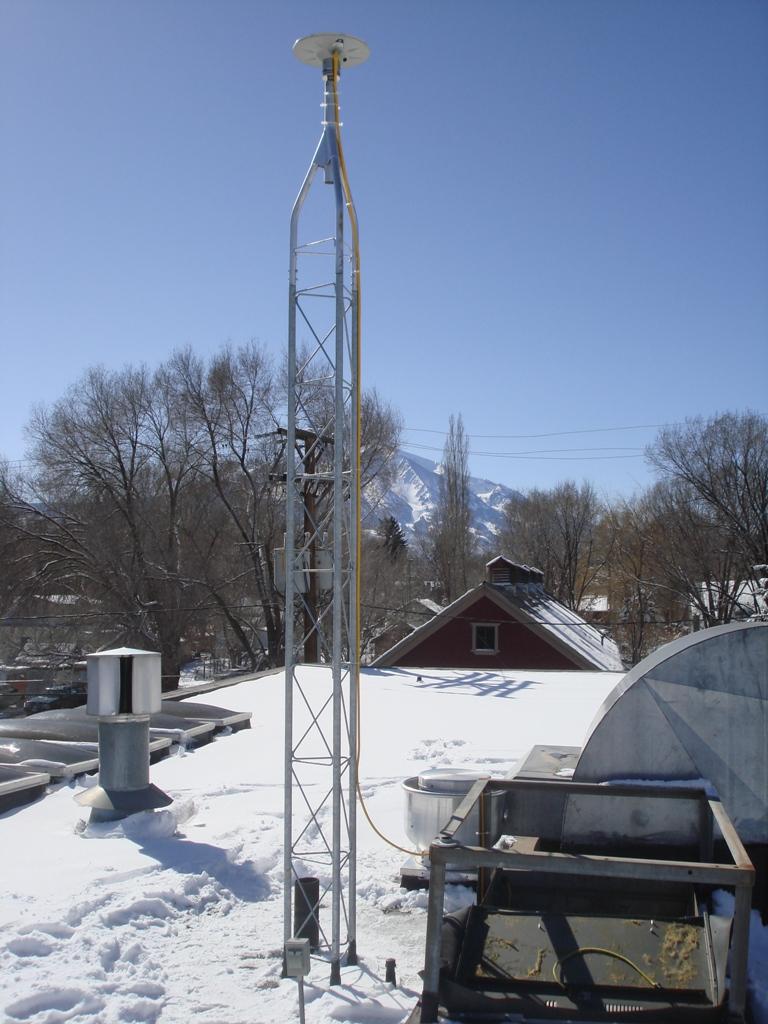 Antenna on a flat, snow-covered roof that has other metal ducts and furnace vent on it with trees, a mountain, and blue sky in the background.