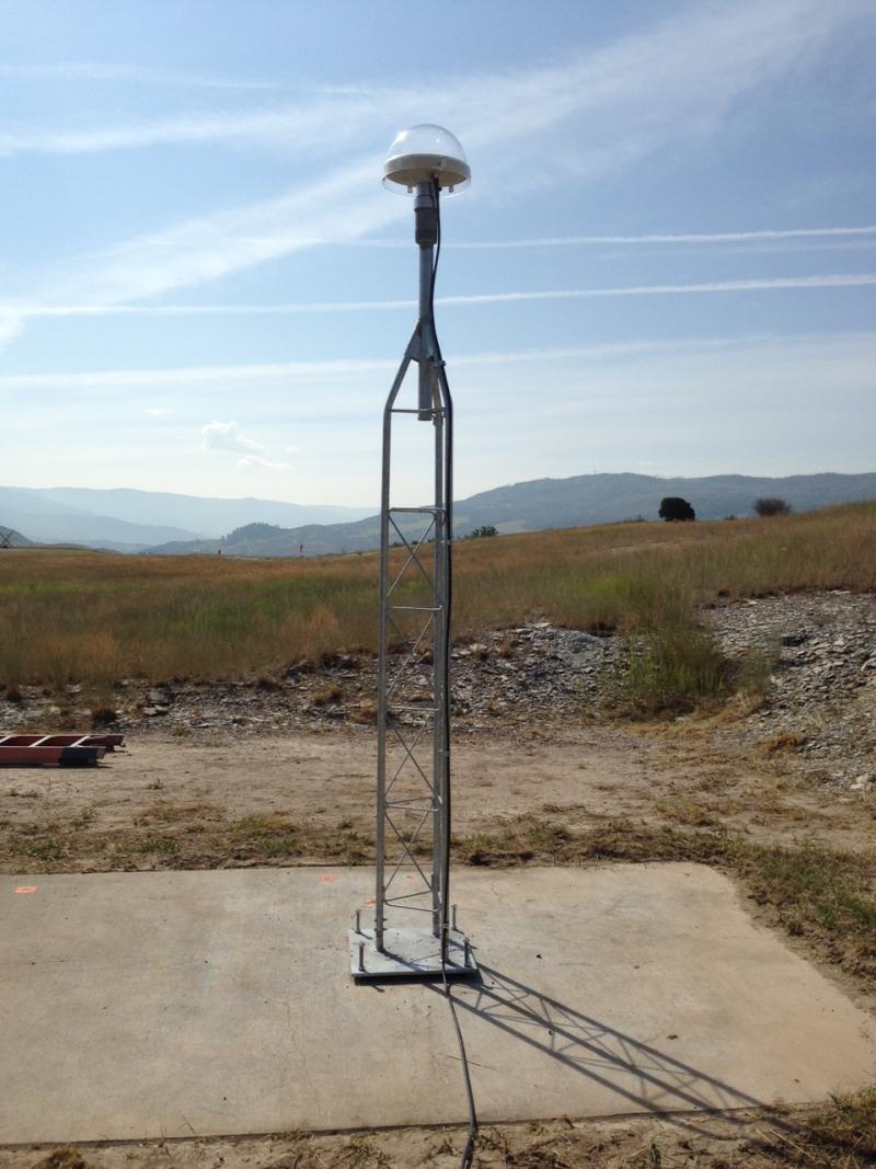 Antenna embedded in a concrete pad surrounded by a grassy area with mountains and an overcast blue sky in the background.