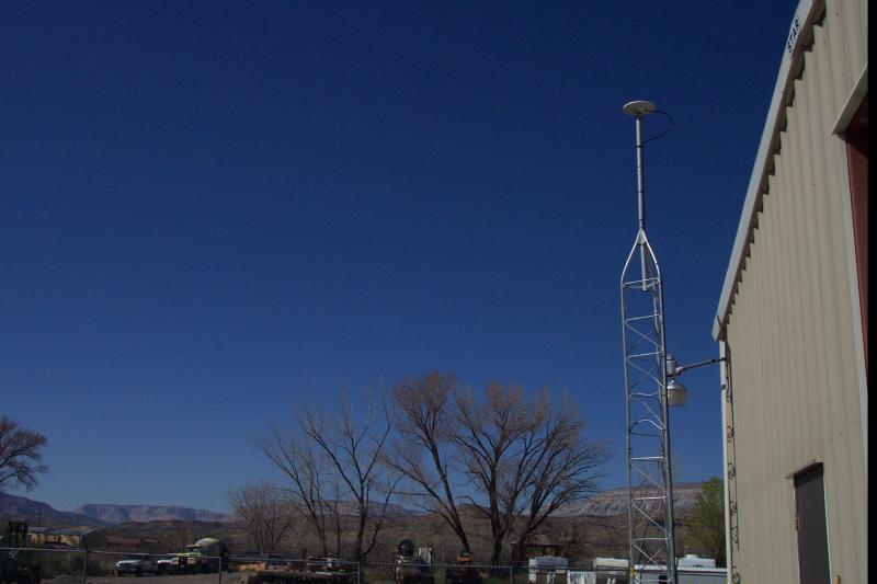 GPS antenna located next to a tan metal building showing road construction vehicles at the building base with mountains and blue sky framing the antenna.