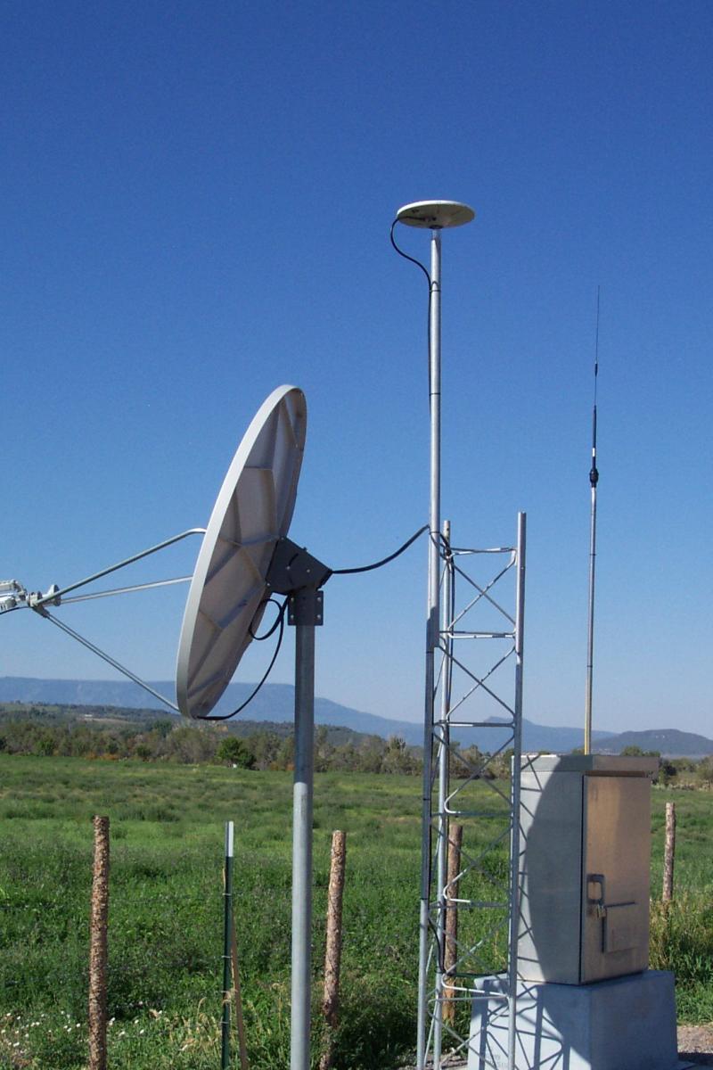 GPS antenna and dish antenna next to a power source on a small gravel plot and next to a green fenced field.