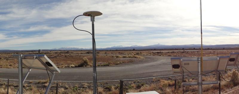 Antenna surrounded by other metal equipment, located in desert area with a U shaped gravel road in front of it, and a blue sky with large wispy clouds.