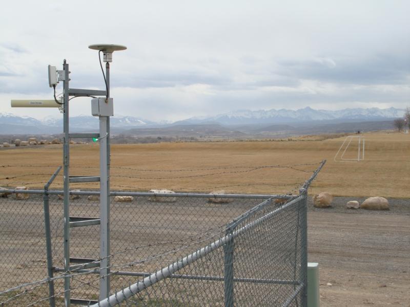 GPS antenna located inside a chain link fence next to a gravel road and a soccer field with a snowy mountainous backdrop.