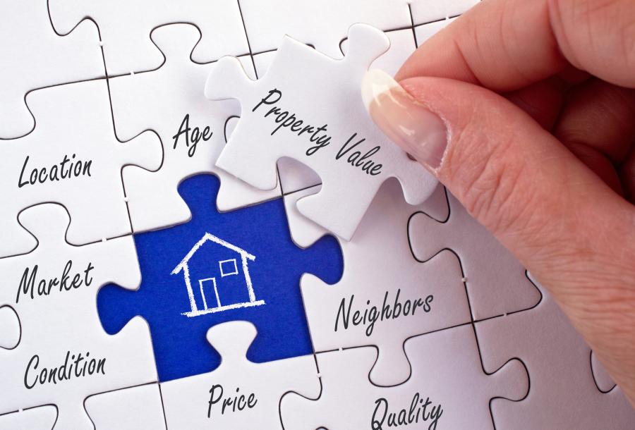 Photograph of jigsaw puzzle with terms written on the pieces for Property Value (this piece is grasped between woman's fingers as she prepares to place it in the puzzle), Location, Age, Market Condition, Neighbors, Price