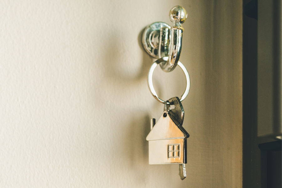 Silver house key with keychain of silver house hang on cream colored wall.